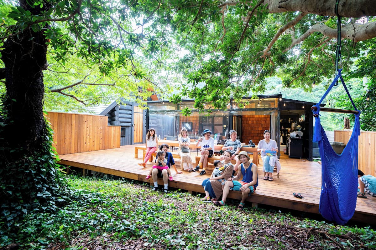 Una terraza de madera con un grupo de personas, una hamaca colgante y un edificio con puerta de cristal.