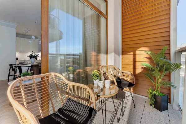 A cozy, covered balcony with two wicker armchairs and a small table set with refreshments. The wooden deck and ceiling panels create a warm, natural atmosphere. Large windows overlook an urban landscape, and a potted plant adds a touch of greenery.