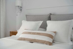 Ein ordentlich bezogenes Bett mit weißer Bettwäsche und zwei Kissen, eines mit Streifenmuster und Schrift, sowie eine wandmontierte Lampe.