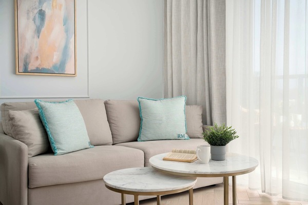 A cozy living area features a plush sofa adorned with light blue pillows and two circular coffee tables. A small potted plant and an abstract art piece add character to the minimalist space.