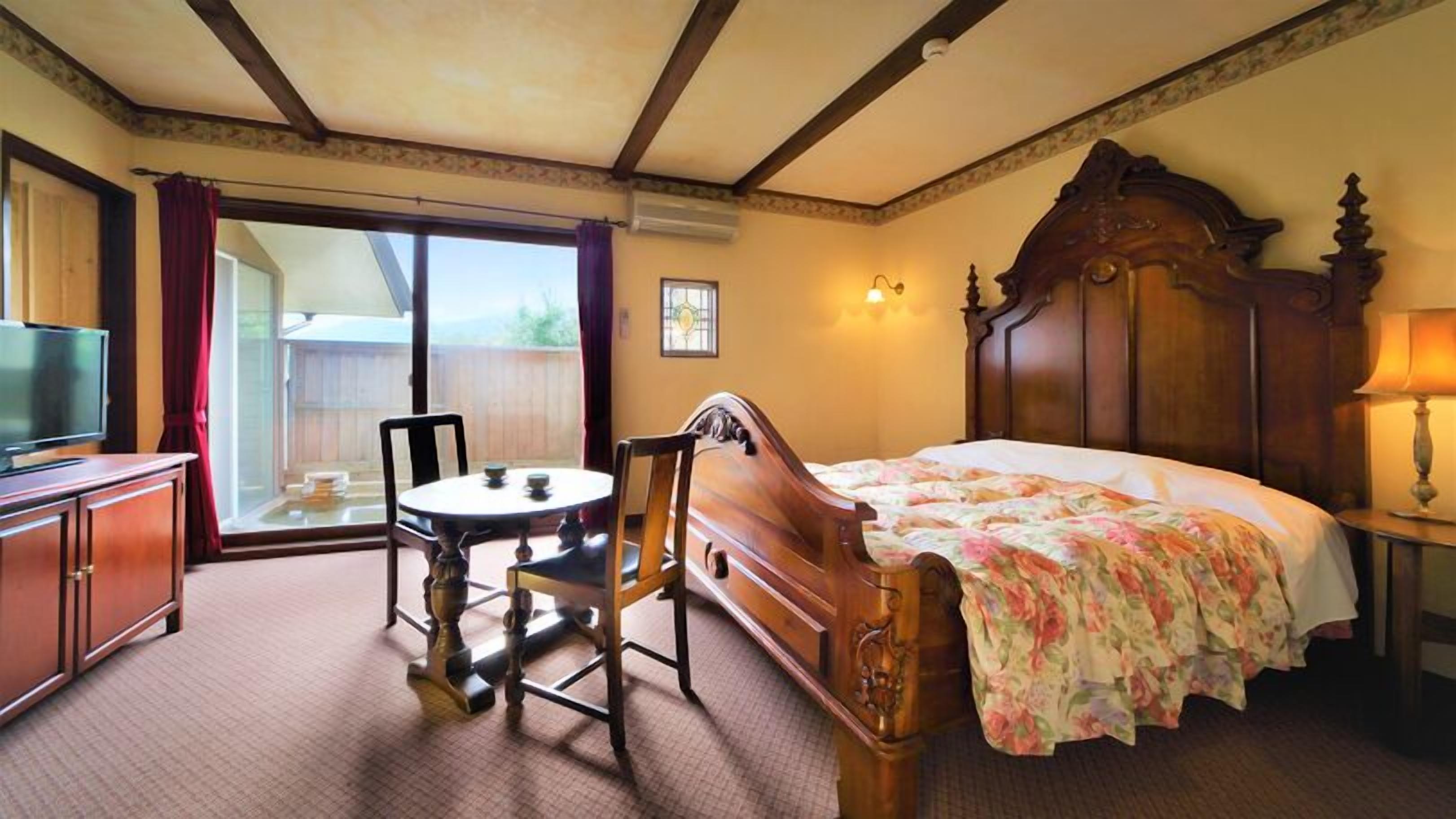 A hotel room with a bed, a television, a table with chairs, and a balcony with a view.