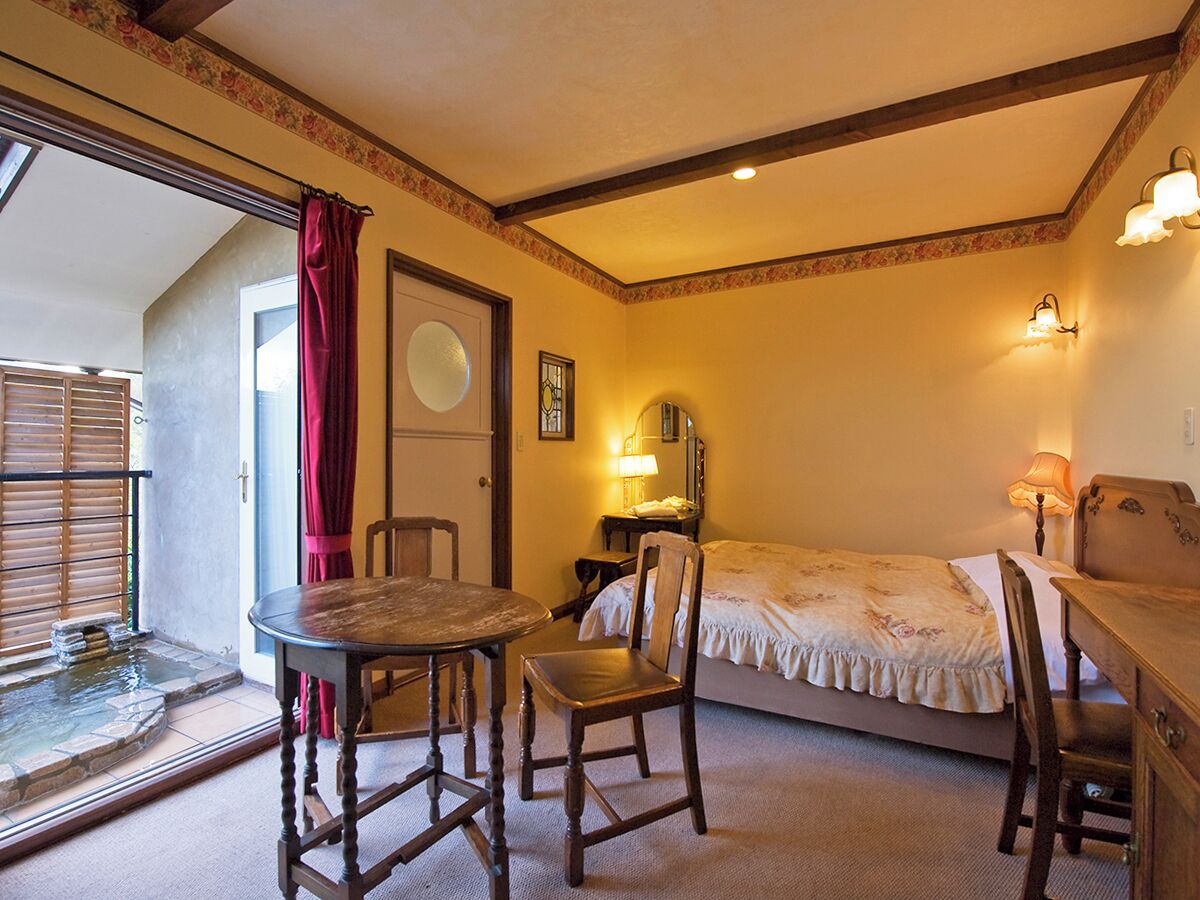 Una habitación con cama, una mesa, sillas, un escritorio, una lámpara, un espejo y una ventana con cortina.