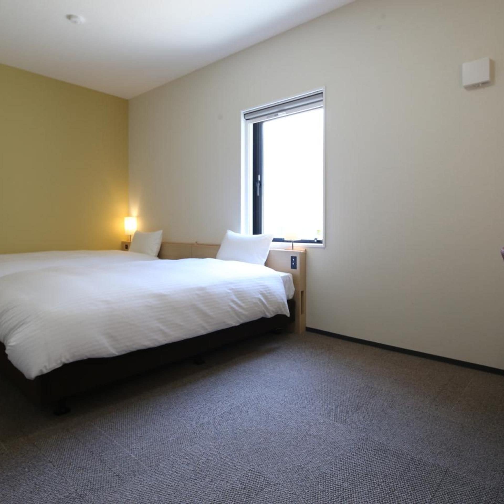 A hotel room with a single bed, a nightstand with a lamp, a window, and a wall-mounted control panel.