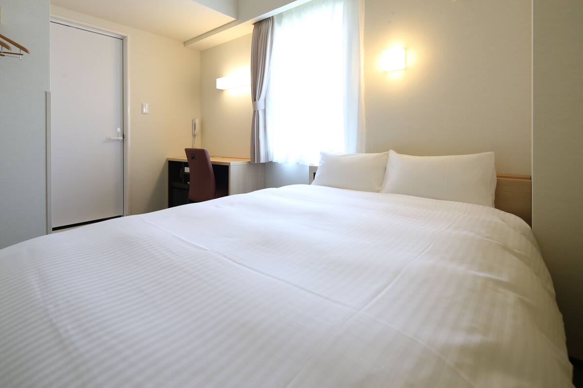 A hotel room with a large bed, a desk, a chair, a wardrobe, and a window with curtains.