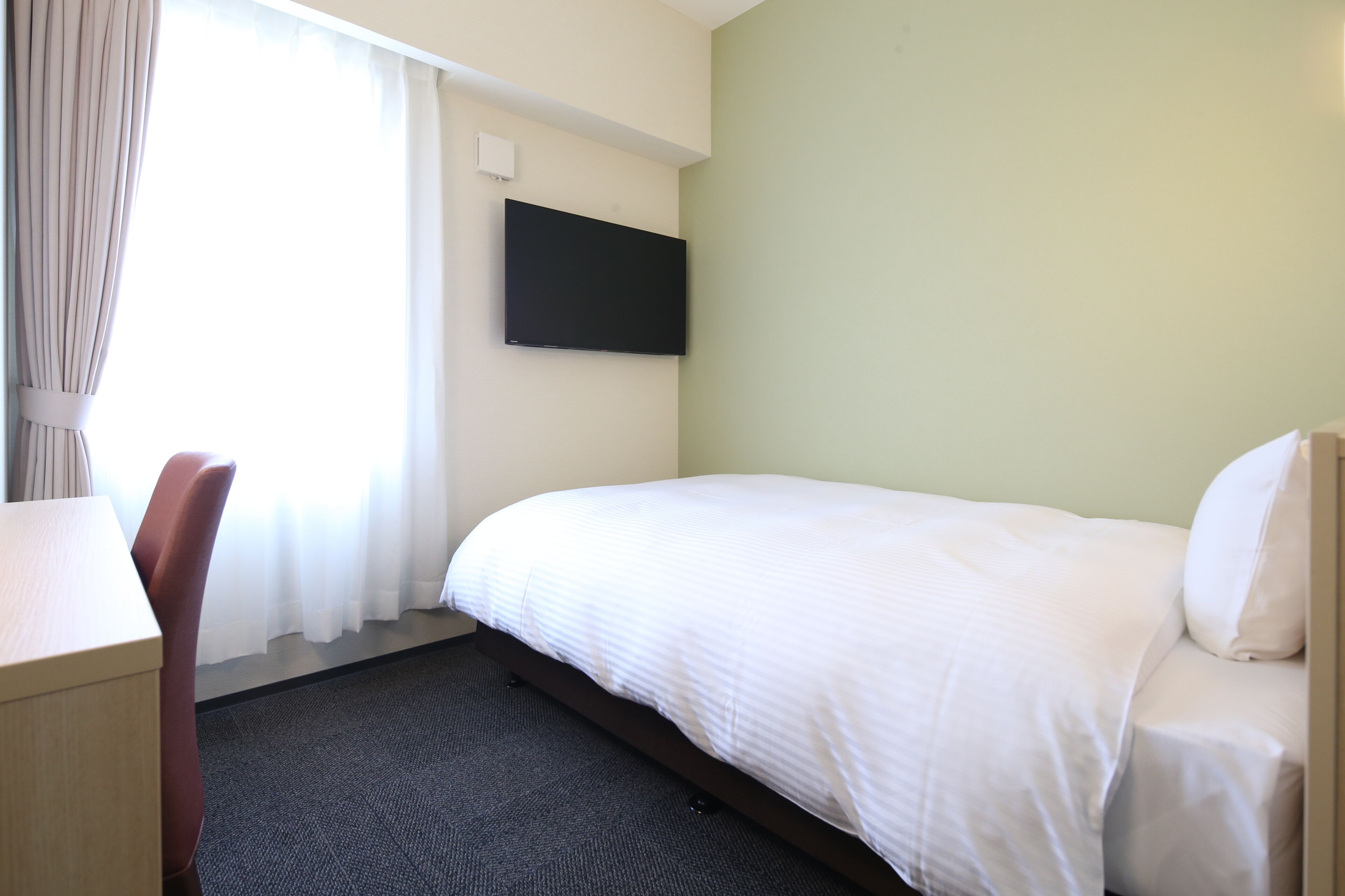 Une chambre d’hôtel équipée d’un lit, d’un bureau, d’une chaise, d’un téléviseur à écran plat fixé au mur et d’une fenêtre avec des rideaux.