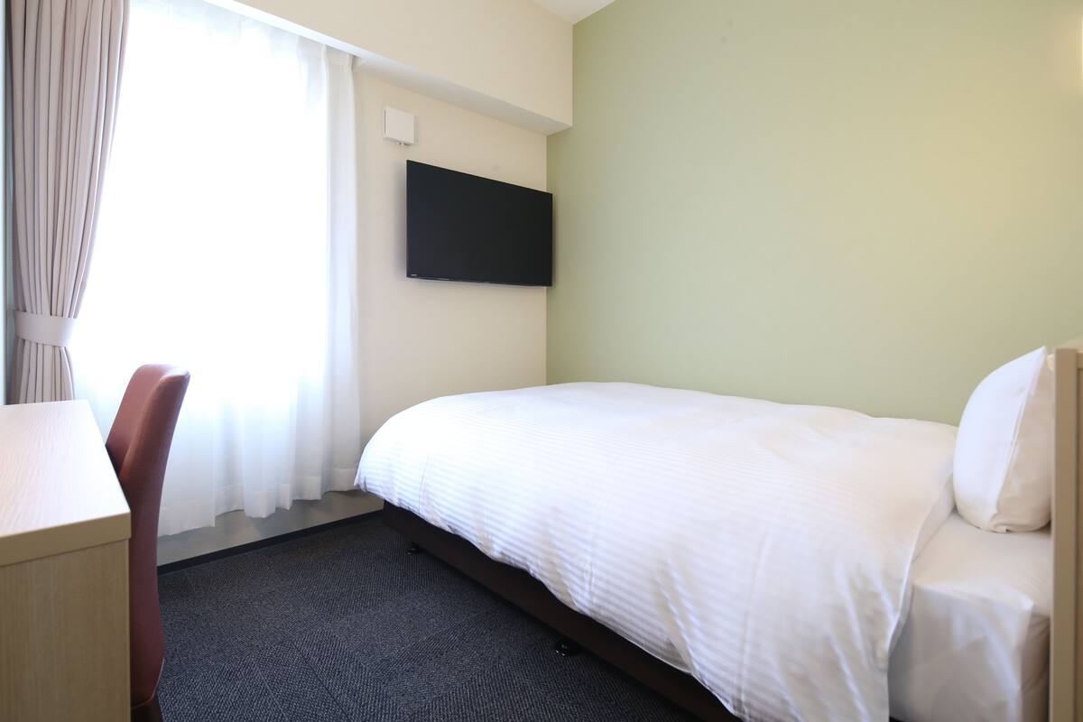 A hotel room with a bed, a desk, a chair, a flat-screen TV mounted on the wall, and a window with curtains.