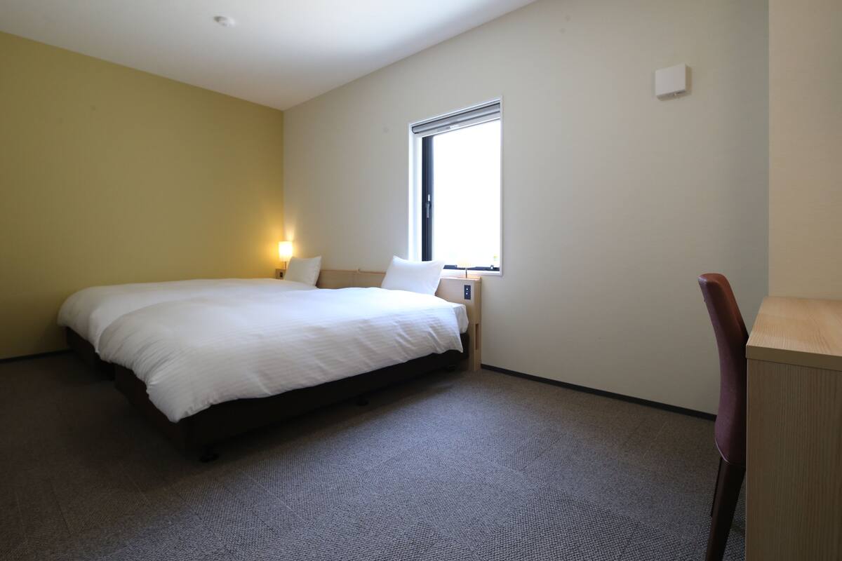 A hotel room with a bed, a desk, a chair, a window, and a wall-mounted light.