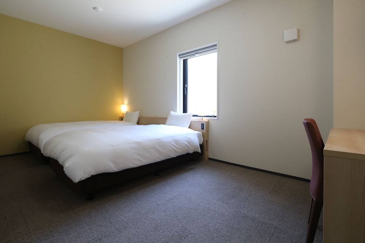 A hotel room with a bed, a nightstand, a chair, a window, and a wall-mounted light.