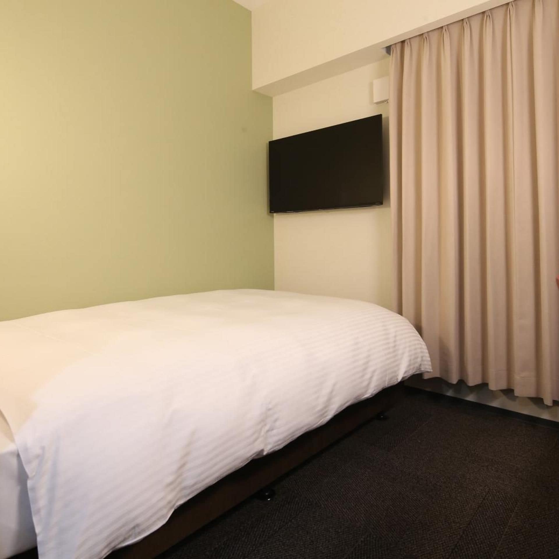A hotel room with a single bed, a flat-screen TV mounted on the wall, and a window with curtains.