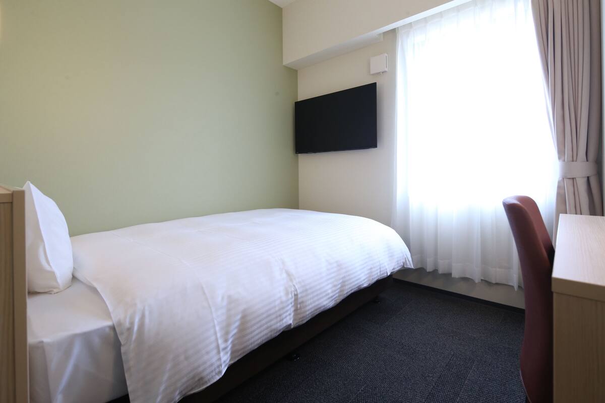A hotel room with a bed, a desk, a chair, a flat-screen TV mounted on the wall, and a window with curtains.