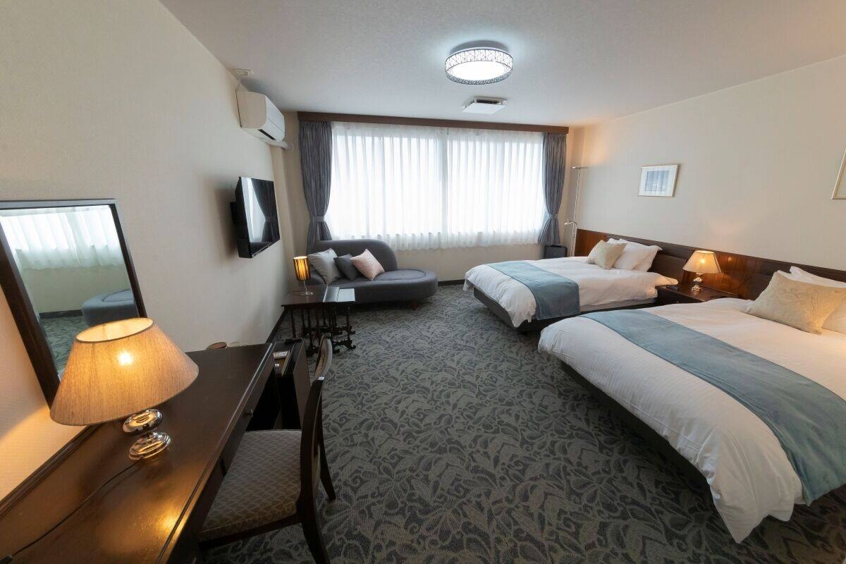 A hotel room with two beds, a desk, a chair, a TV, and a large window with curtains.