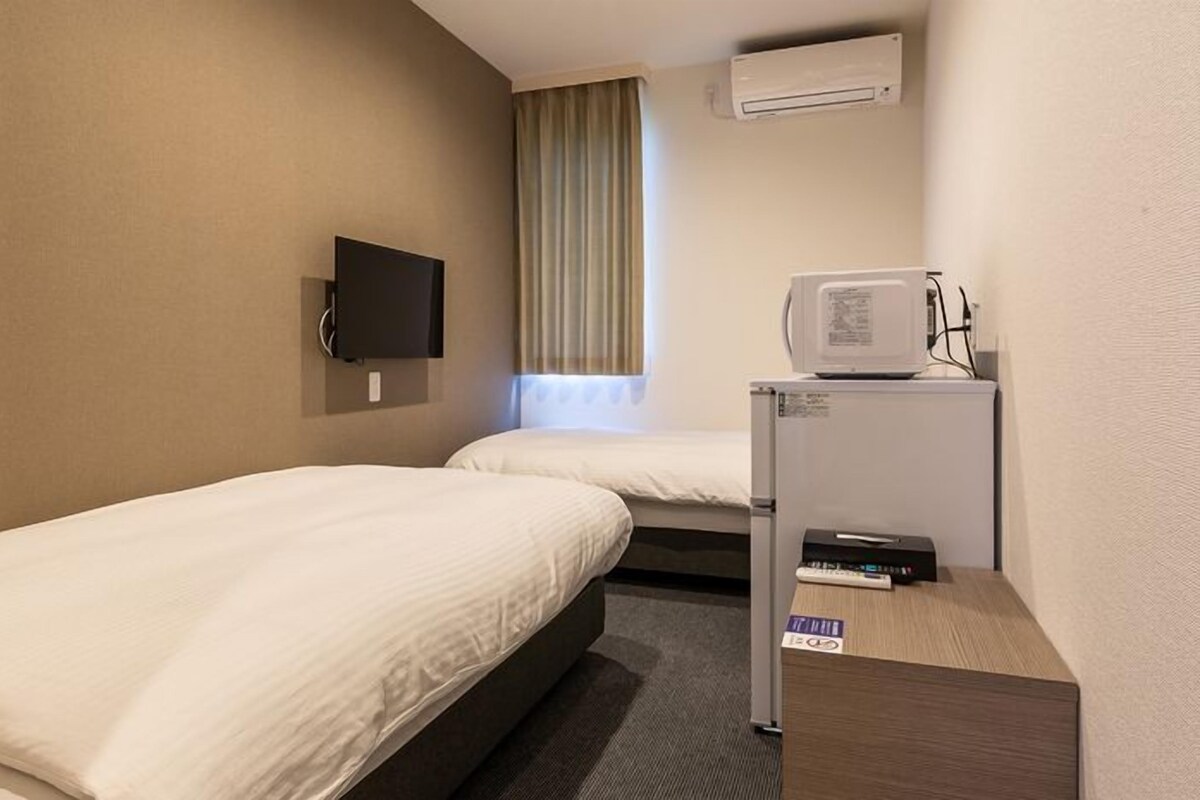 A hotel room with two single beds, a TV mounted on the wall, an air conditioning unit, and a small desk with a printer.