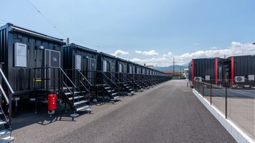 A row of industrial-style buildings with numbered doors, a gravel ground, and a clear sky.