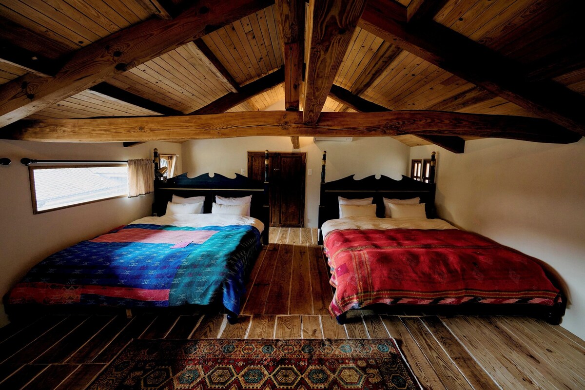 Een kamer met twee bedden, houten balken in het plafond, een groot raam en een kamerbreed tapijt met dessin.