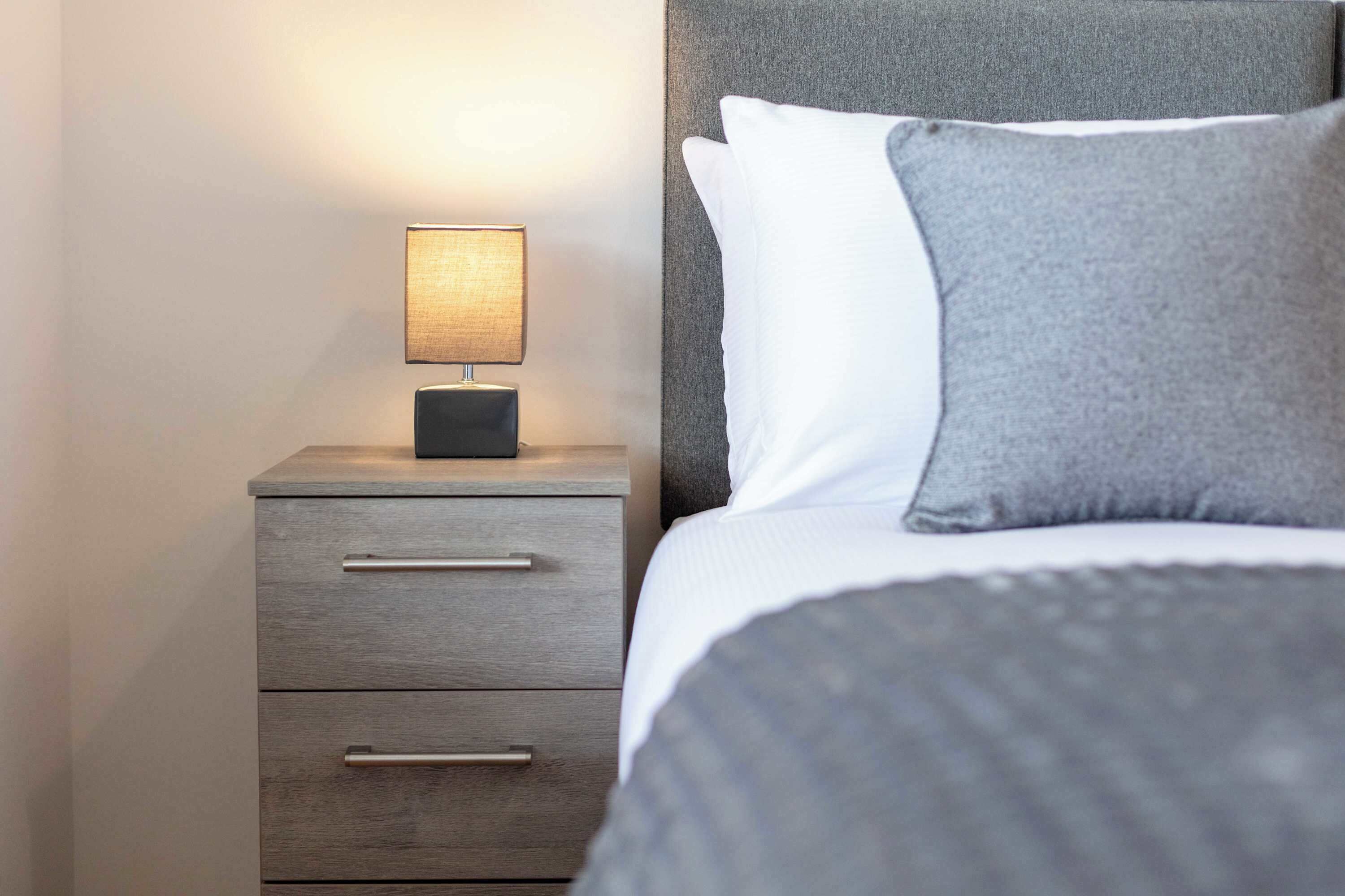 A neatly made bed with a grey headboard, a bedside lamp, and a wooden nightstand with drawers.