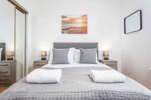 Uma cama bem arrumada com almofadas, uma mesa de cabeceira com abajur, um quadro na parede e um espelho.