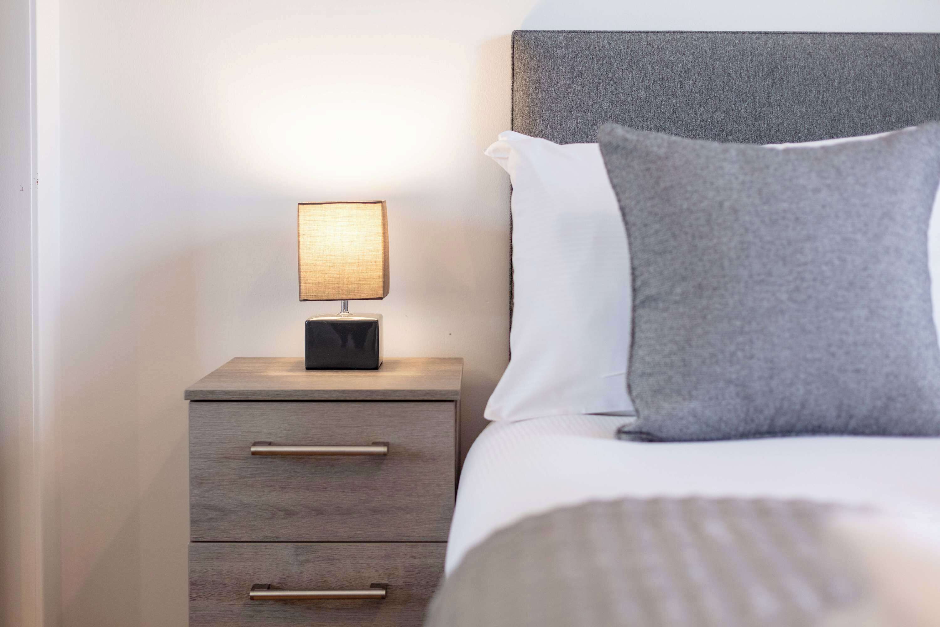A neatly made bed with a grey pillow, a bedside lamp, and a wooden nightstand with drawers.
