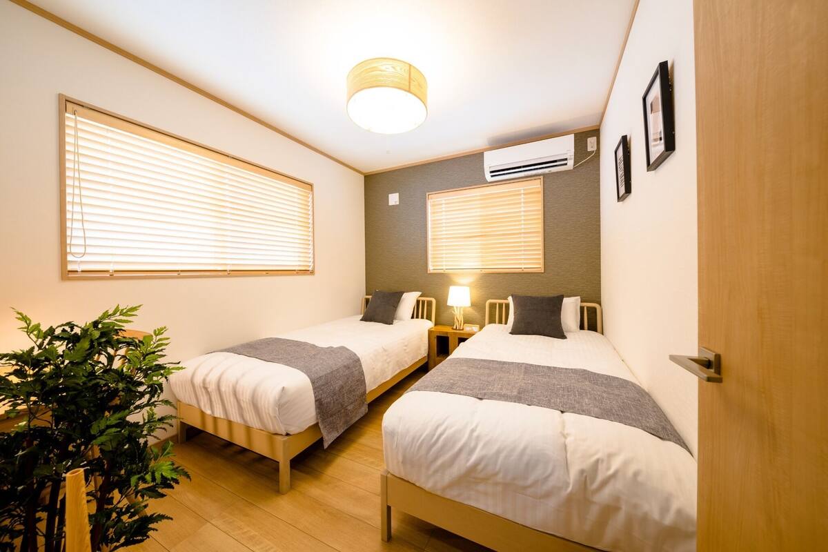 A hotel room with two single beds, a bedside table with a lamp, a potted plant, and an air conditioning unit on the wall.