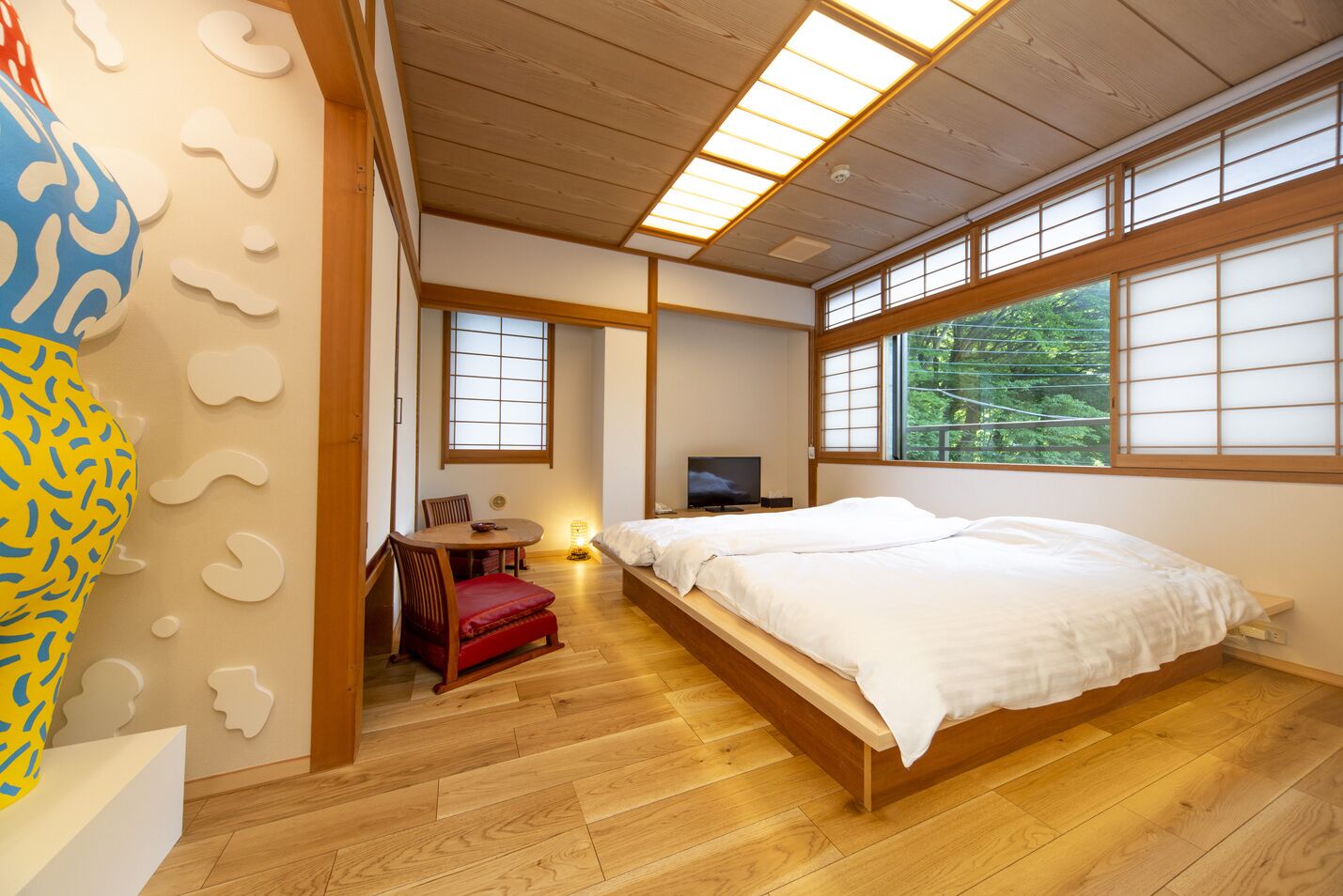 Un dormitorio tradicional japonés con suelo de tatami, una cama baja de madera, un televisor y vistas a la vegetación exterior.