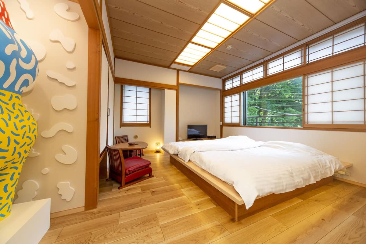 Un dormitorio tradicional japonés con suelo de tatami, una cama baja de madera, un televisor y vistas a la vegetación exterior.