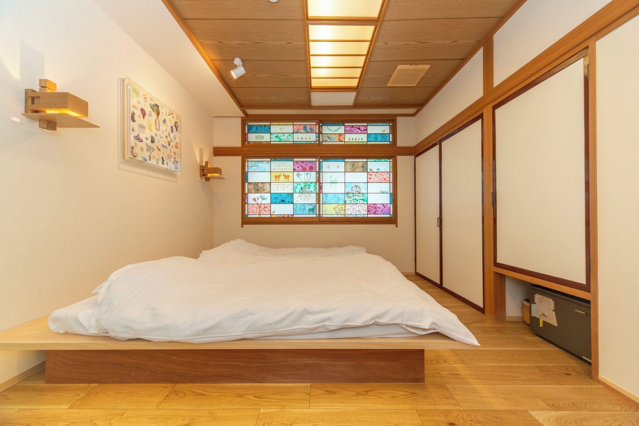 Una habitación con cama, piso de madera, techo con iluminación empotrada y ventanal con vidrieras de colores, además de puertas correderas.