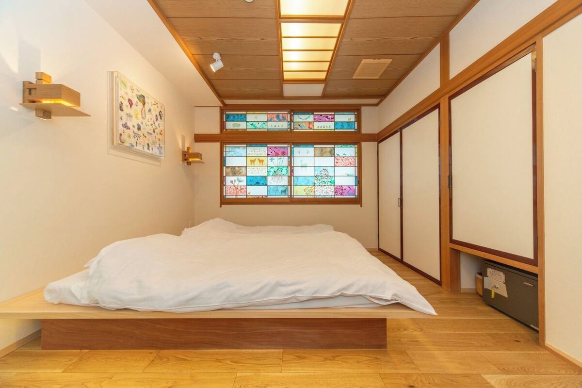 Una habitación con cama, piso de madera, techo con iluminación empotrada y ventanal con vidrieras de colores, además de puertas correderas.
