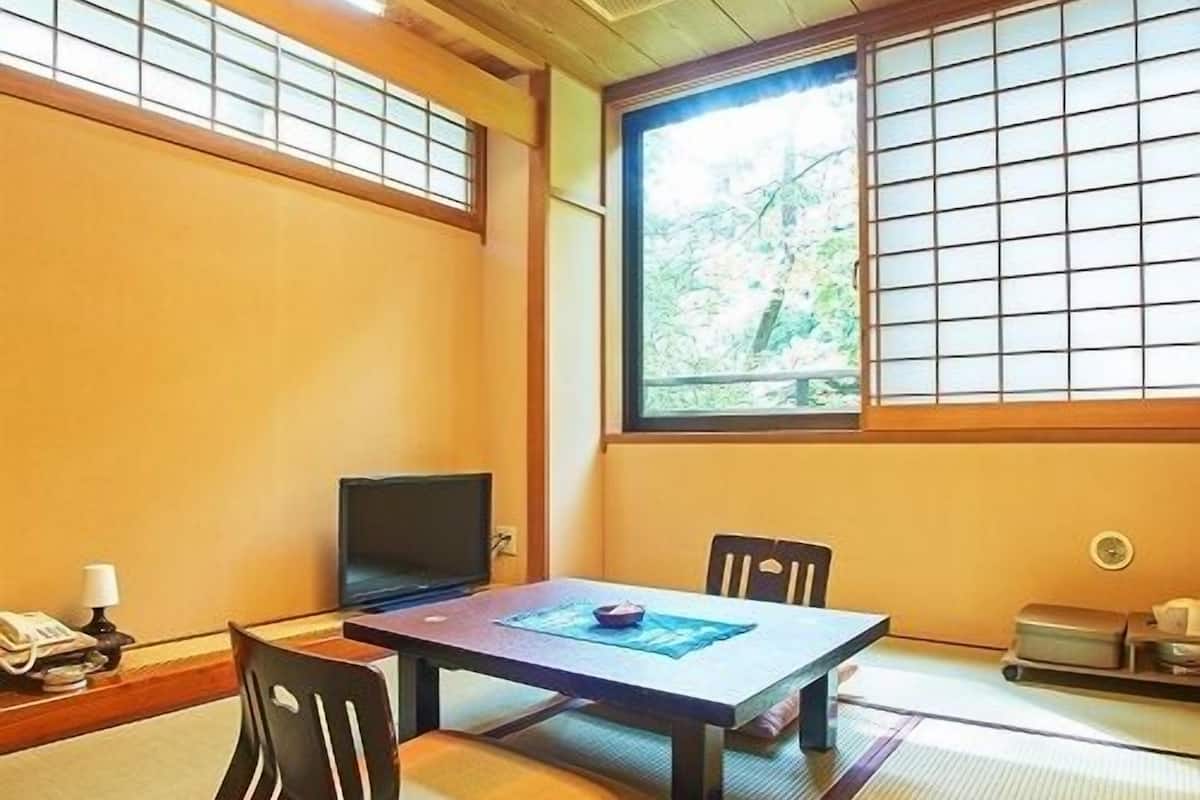 Una habitación japonesa tradicional con suelo de tatami, una mesa baja, un televisor y una ventana con vistas a árboles.