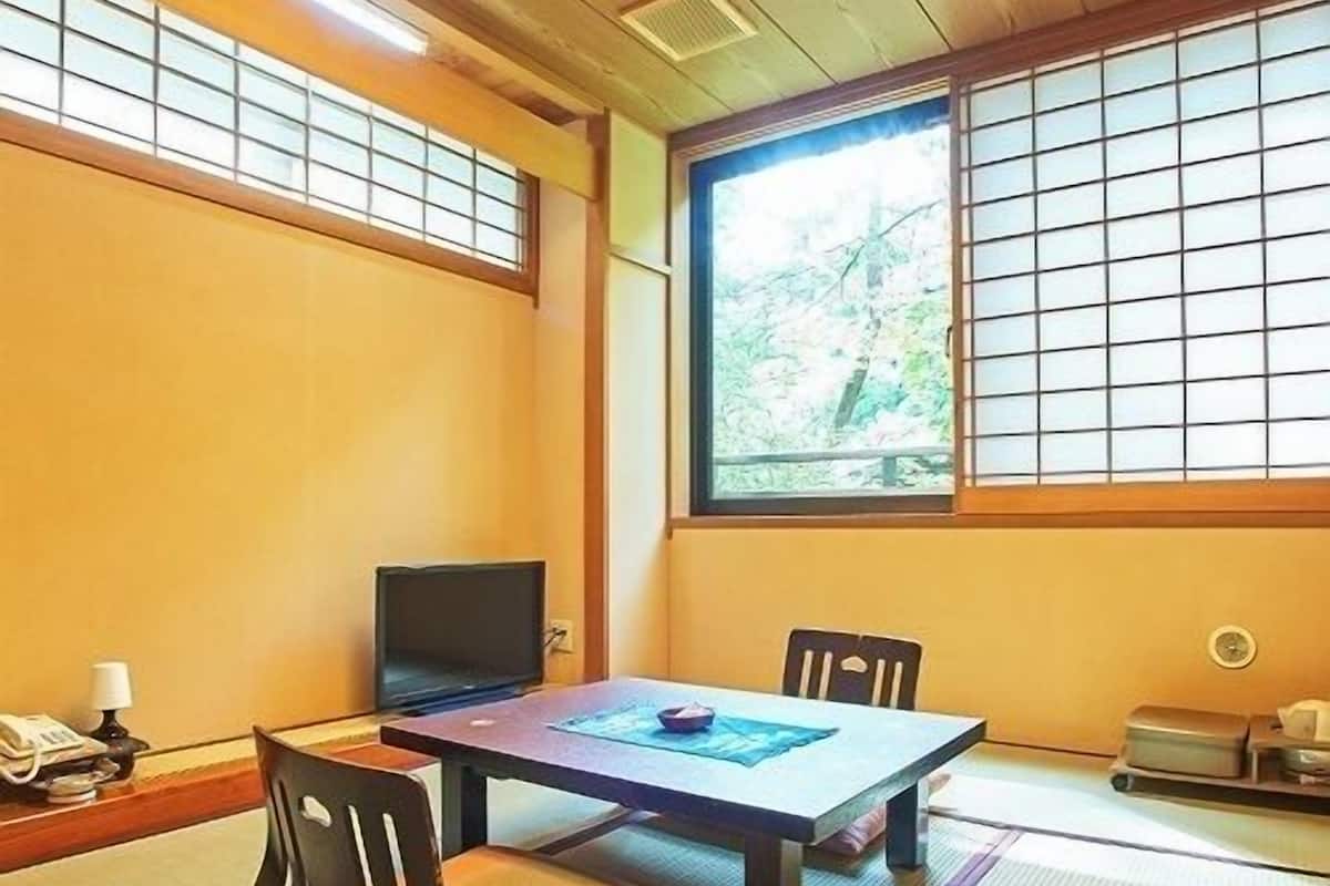 Una habitación tradicional japonesa con suelo de tatami, una mesa baja, un televisor y una ventana con vistas a árboles.