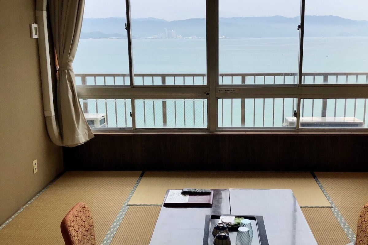 Une pièce avec un sol recouvert de tatamis, une table avec une télécommande et une vue sur la mer grâce à de grandes fenêtres.
