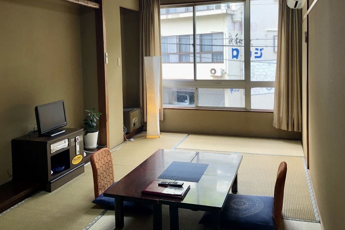 Une pièce avec un sol recouvert de tatamis, une table basse avec des coussins, un téléviseur posé sur un meuble et une fenêtre donnant sur des immeubles.