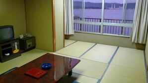 Une pièce avec un sol recouvert de tatamis, une télévision, une petite table sur laquelle repose un livre rouge, et une fenêtre donnant sur un plan d’eau.