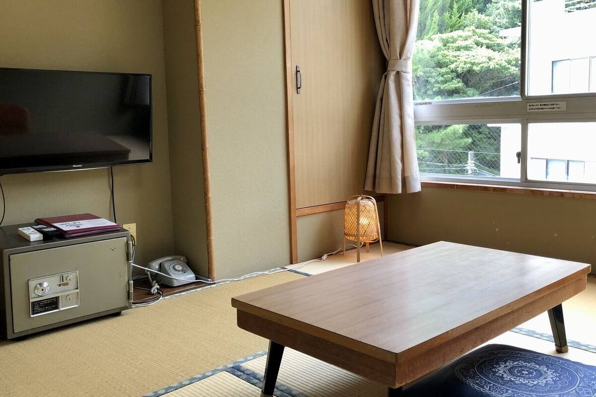 Une pièce équipée d’une télévision, d’une petite armoire, d’une table en bois, d’un tatami japonais traditionnel et d’une fenêtre donnant sur un paysage verdoyant.