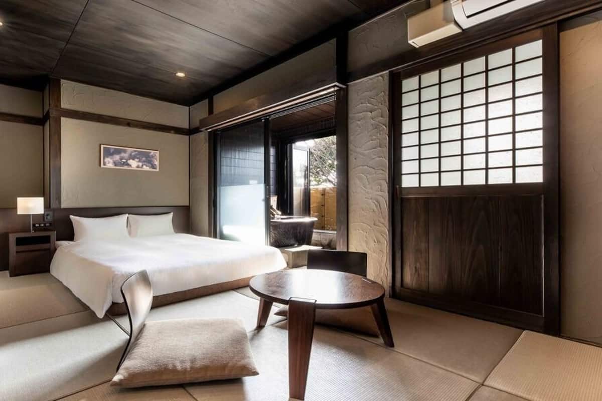 A traditional Japanese-style room with tatami flooring, a bed, a small table, and a window with a grid pattern.