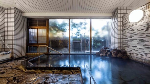 A hot spring area with stone flooring, a wooden bench, and a view of trees through large glass doors. - Nagomi Tsuki (Beppu)