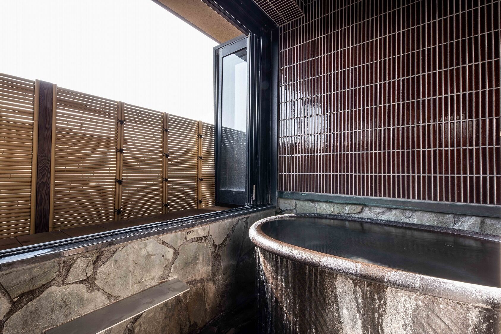 A stone-walled hot tub area with a bamboo fence and a large window.