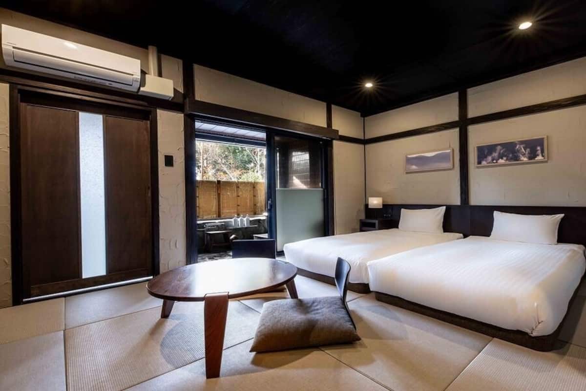 A hotel room with two beds, a dining table, and a sliding door leading to an outdoor area.