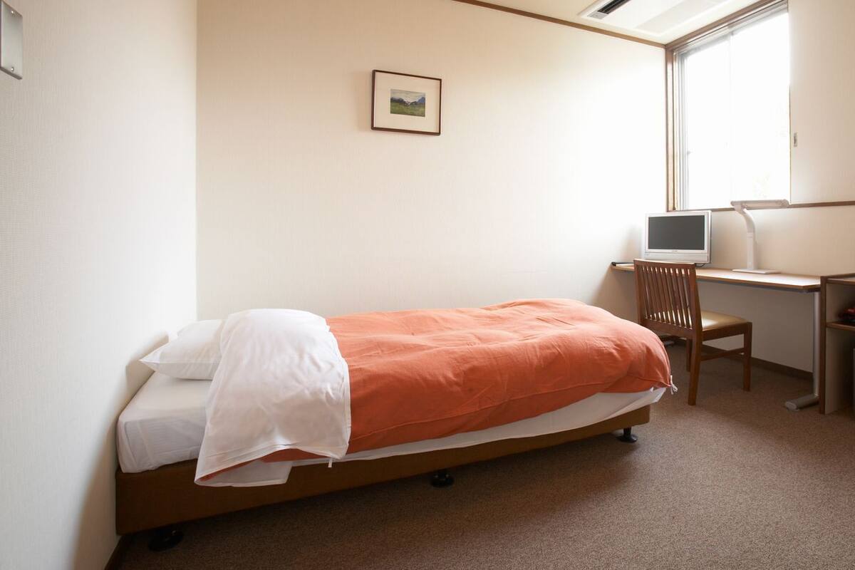 Ein Schlafzimmer mit einem Bett, einem Schreibtisch mit Computer, einem Stuhl, einem Fenster und einem kleinen Safe.