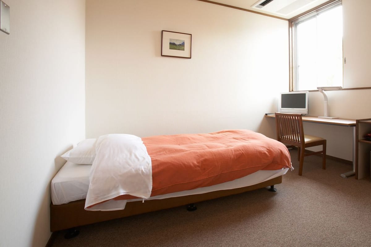 Une chambre à coucher comprenant un lit, un bureau avec un ordinateur, une chaise, une fenêtre et un petit coffre-fort.