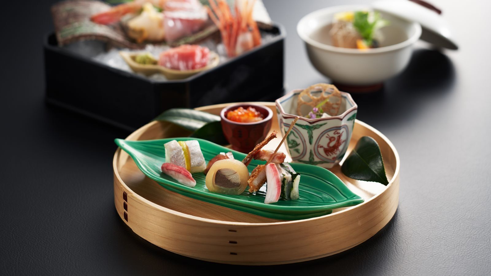 A bamboo steamer with sushi on a green leaf, a bowl of soup, and a small dish of sauce.