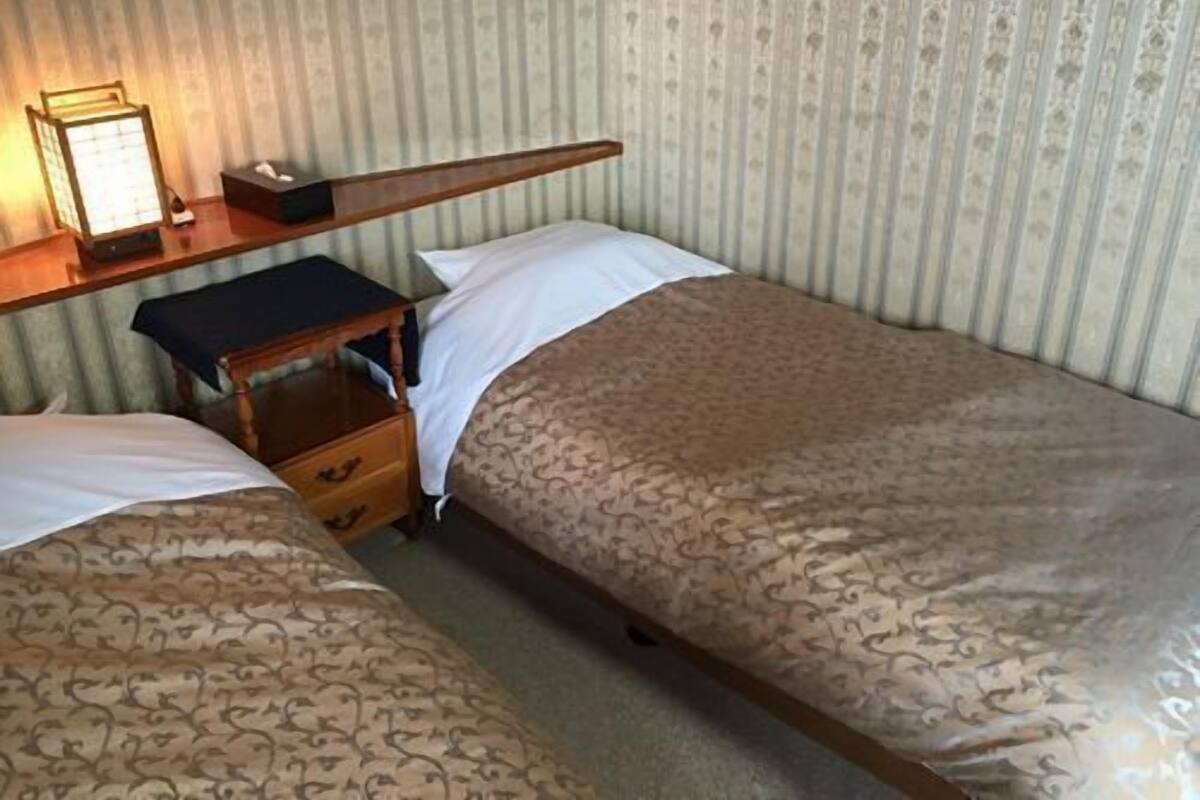 Ein Einzelbett mit einem gemusterten Bettüberwurf, ein Nachttisch mit Lampe sowie eine kleine Kommode.