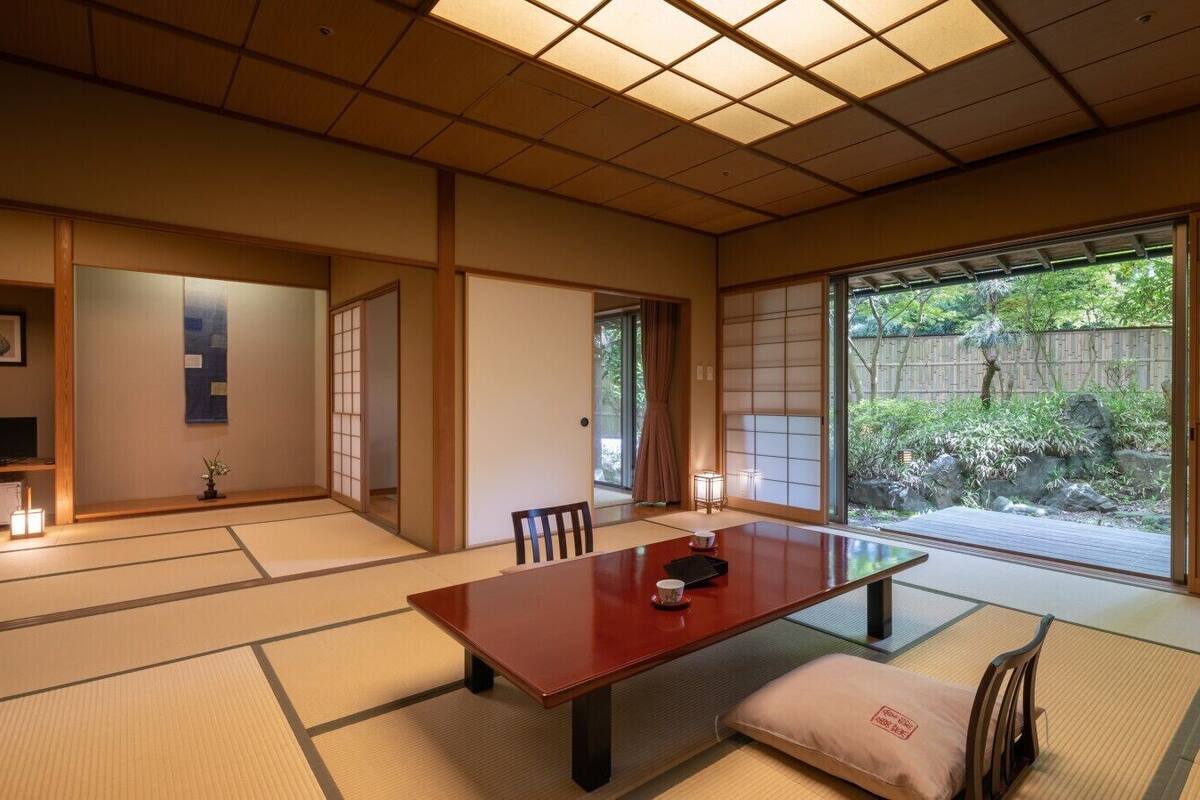 Ein traditioneller japanischer Raum mit Tatami-Böden, einem niedrigen Tisch, Schiebetüren und Blick auf einen Garten.