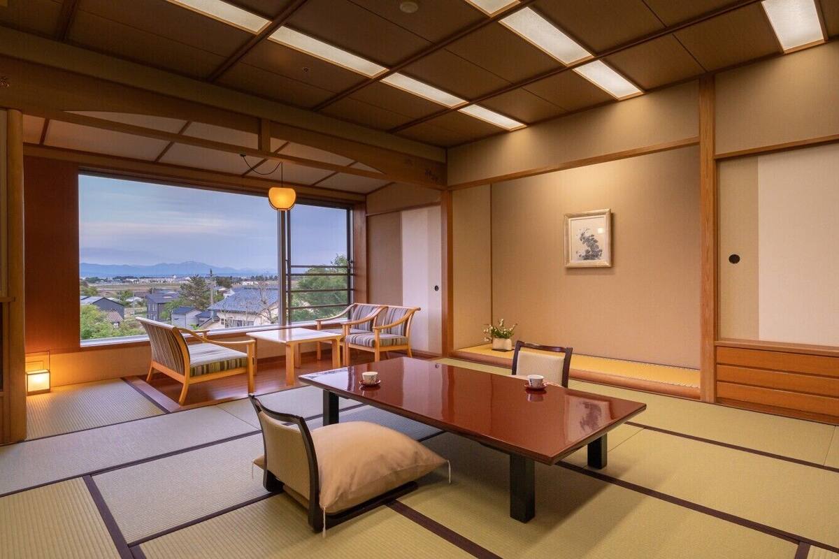Ein traditioneller japanischer Raum mit Tatami-Böden, einem niedrigen Tisch, Stühlen, Blick durch ein großes Fenster und einer Decke mit Holzbalken und eingelassener Beleuchtung.