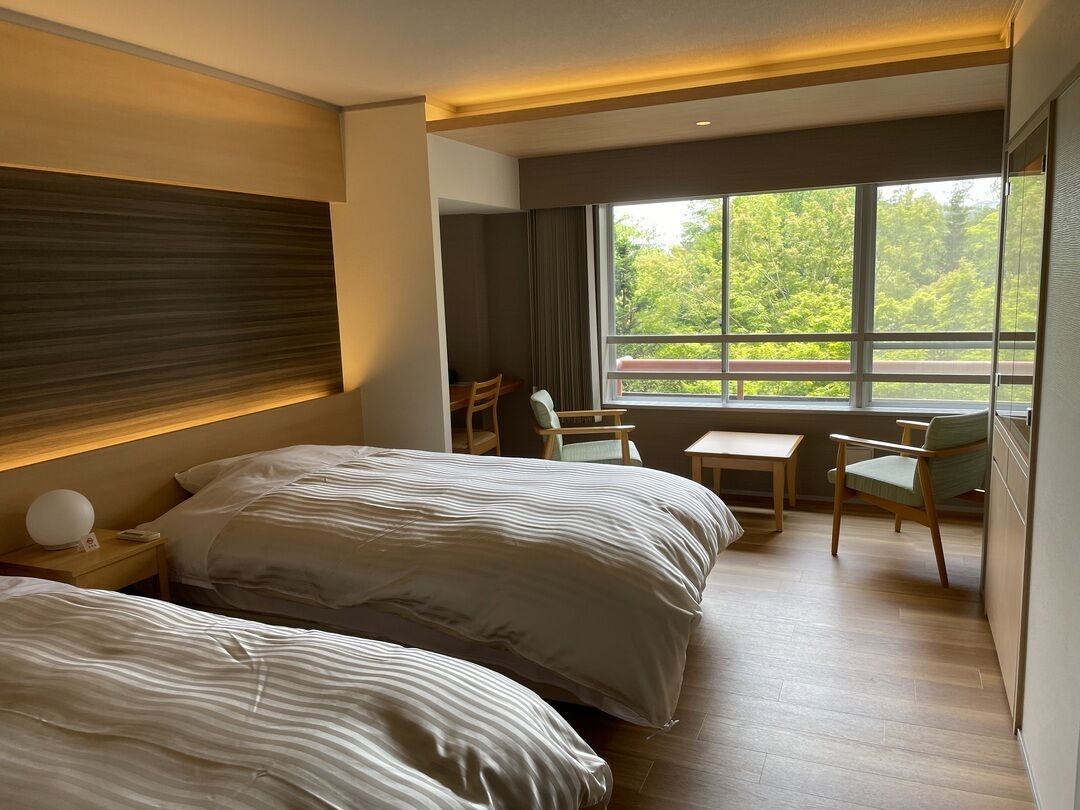 A hotel room with two beds, a small table, chairs, a nightstand, and a large window with a view of greenery.