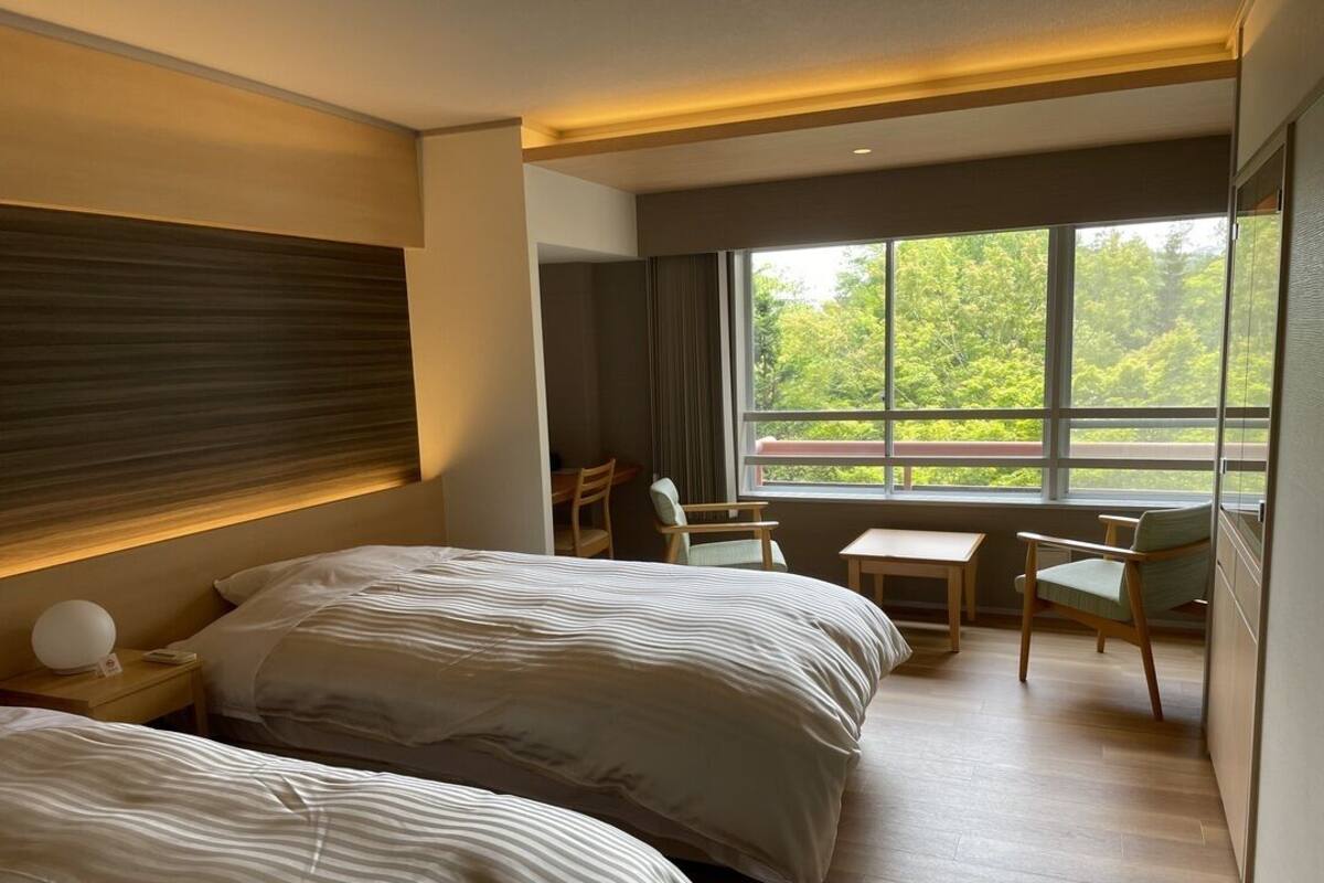 Ein Hotelzimmer mit zwei Betten, einem kleinen Tisch, Stühlen, einem Nachttisch und einem großen Fenster mit Blick auf Grünflächen.