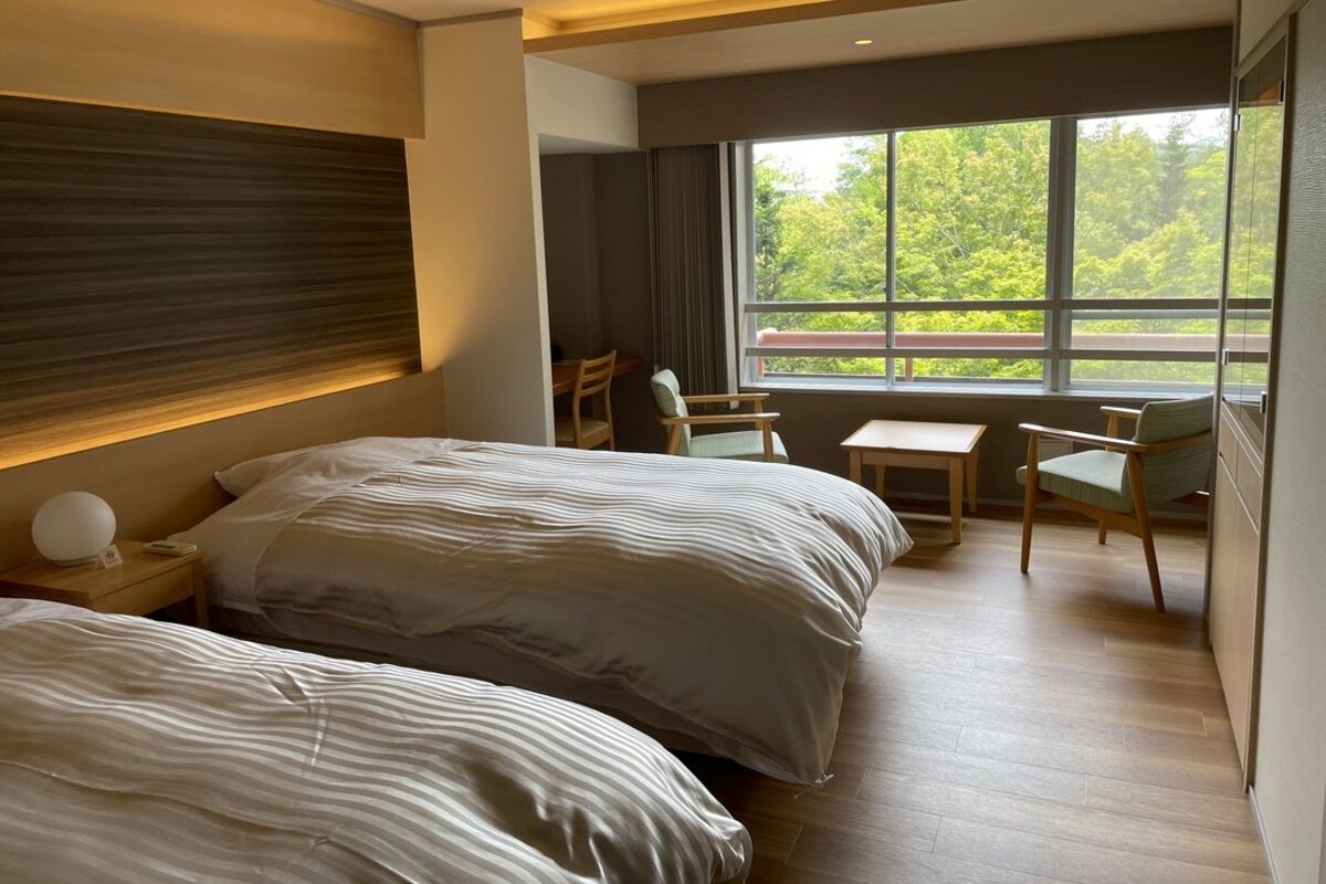 A hotel room with two beds, a small table, chairs, a nightstand, and a large window with a view of greenery.