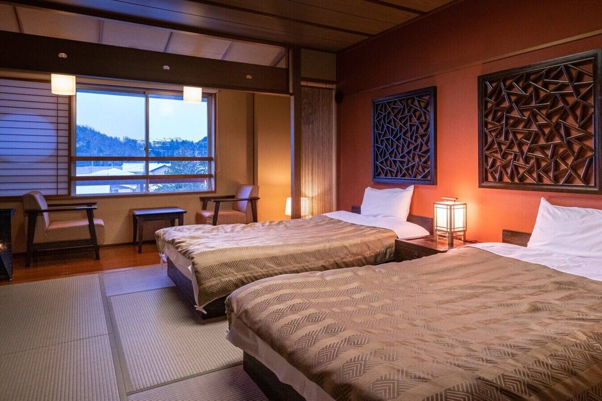 Ein Hotelzimmer mit zwei Betten, einer Sitzecke mit Sessel und Tisch sowie einem Fenster mit Blick auf eine verschneite Landschaft.