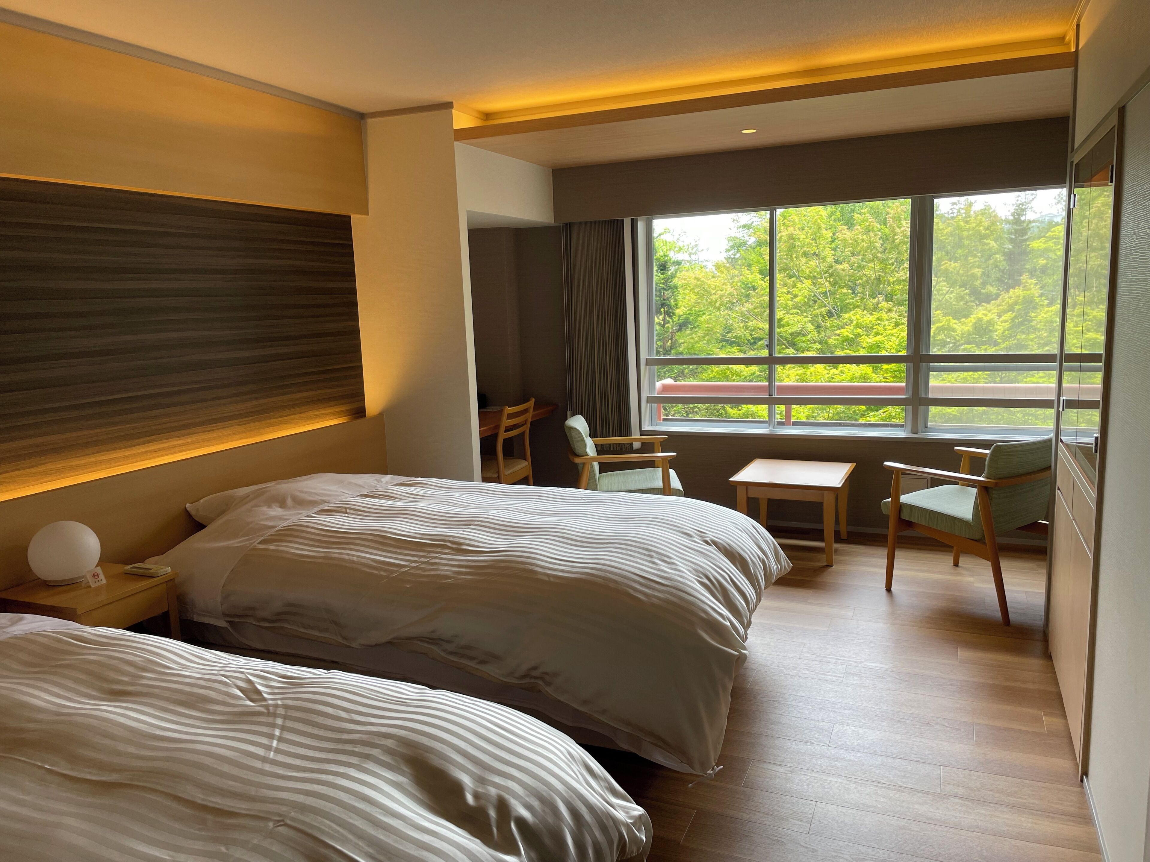 Une chambre d’hôtel avec deux lits, un bureau, une chaise et une grande fenêtre donnant sur la forêt.