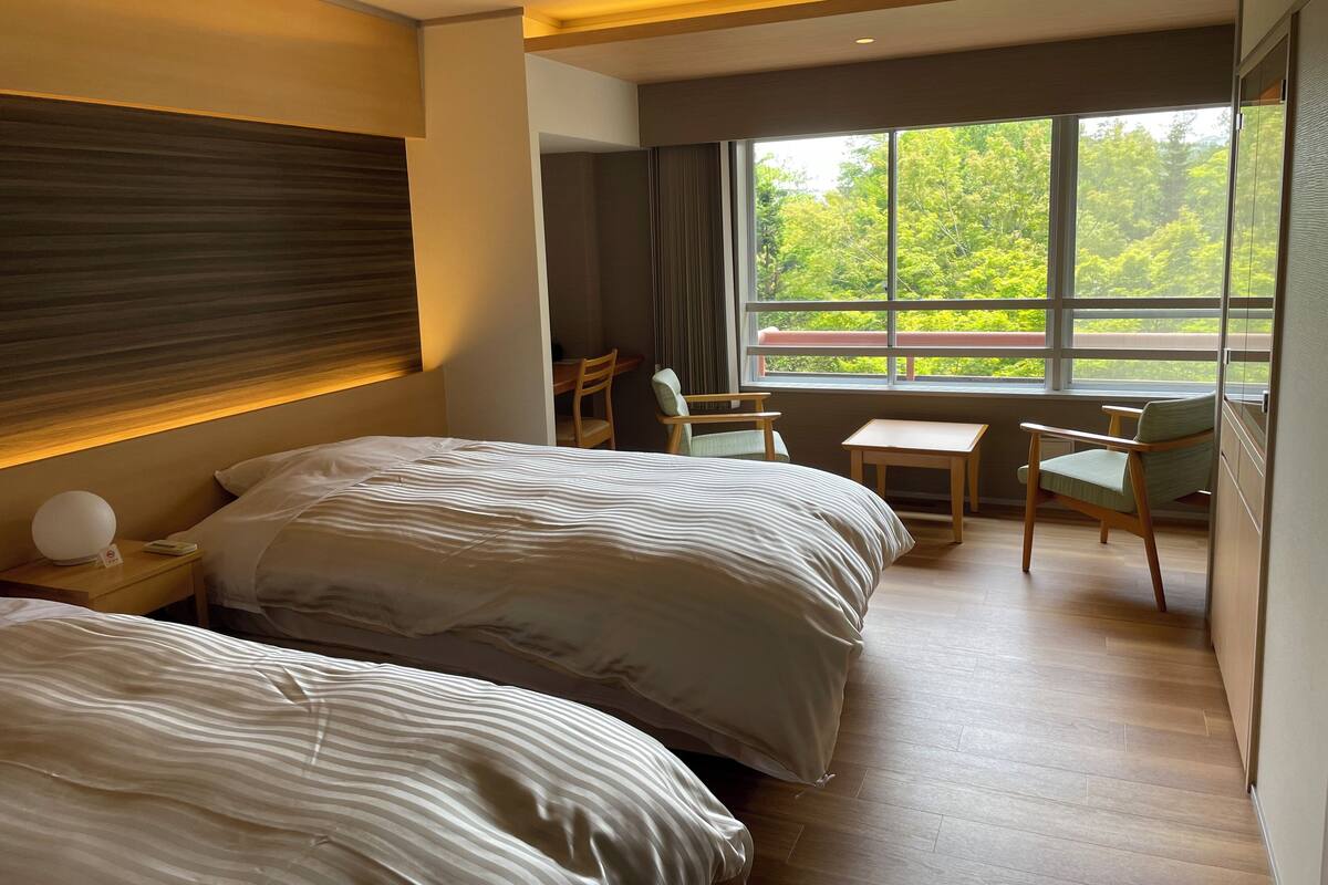 Ein Hotelzimmer mit zwei Betten, einem Schreibtisch, einem Stuhl und einem großen Fenster mit Blick auf Bäume.