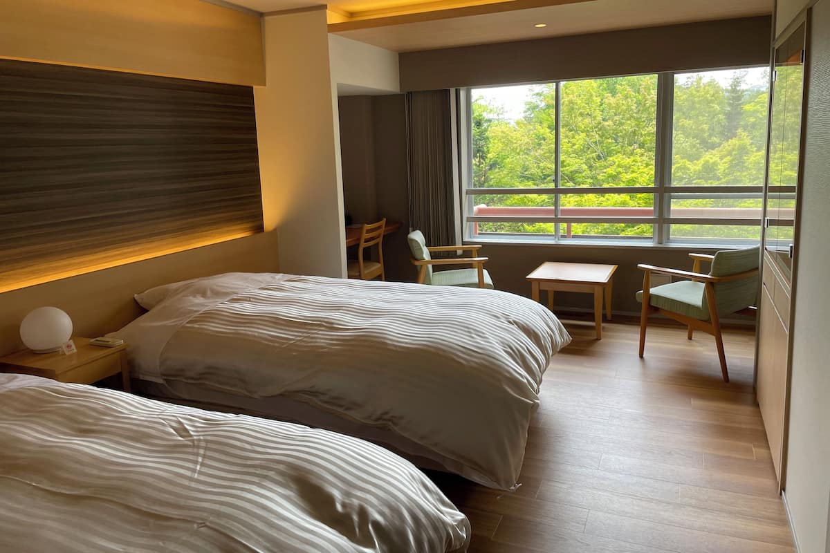 Une chambre d’hôtel avec deux lits, un bureau, une chaise et une grande fenêtre donnant sur la forêt.