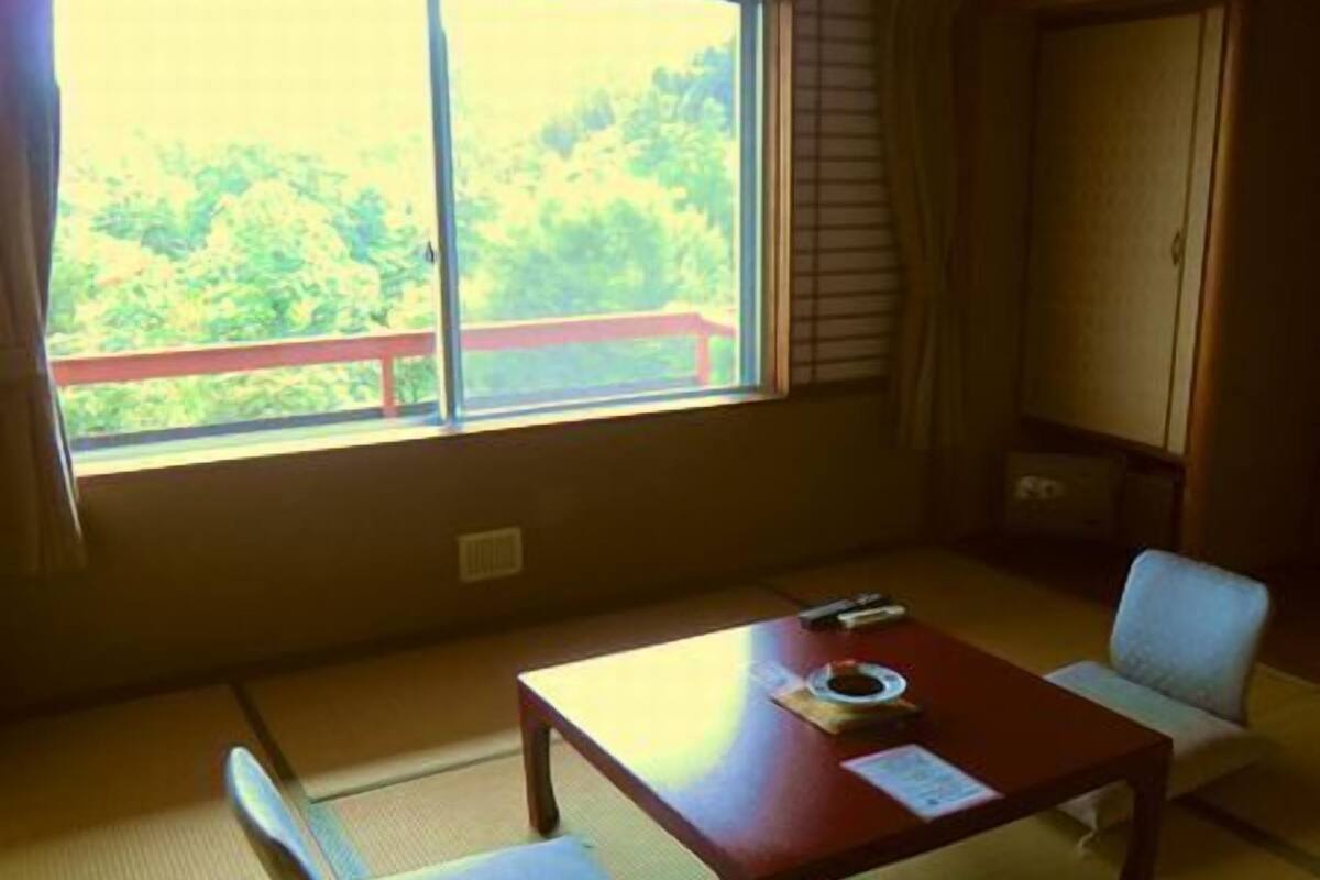 Ein Zimmer mit Tatami-Boden, einem niedrigen Tisch, einem Stuhl und Blick auf Grün durch ein Fenster.